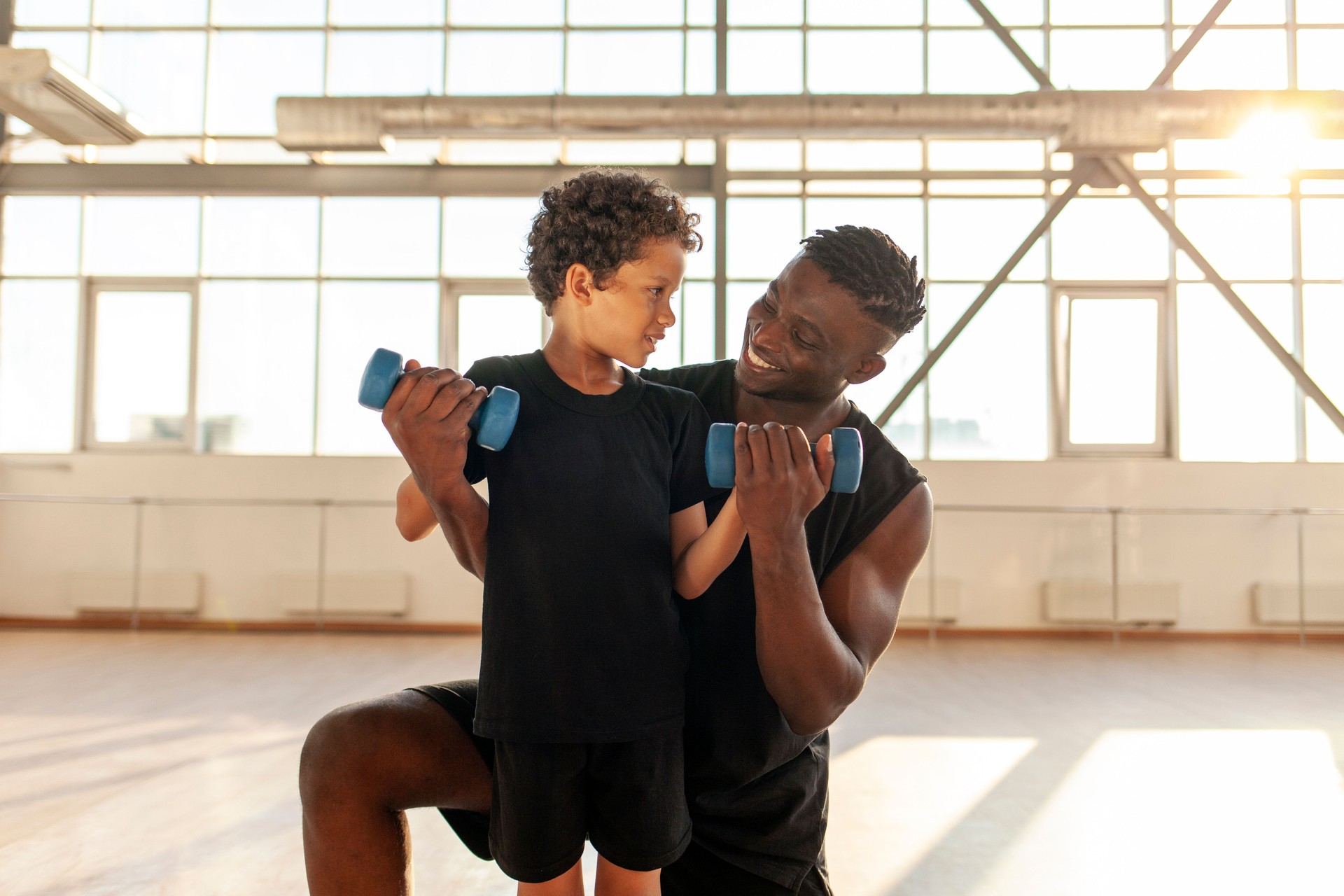 African American boy working out and lifting dumbbells in the gym with his dad, man training his son and helping him